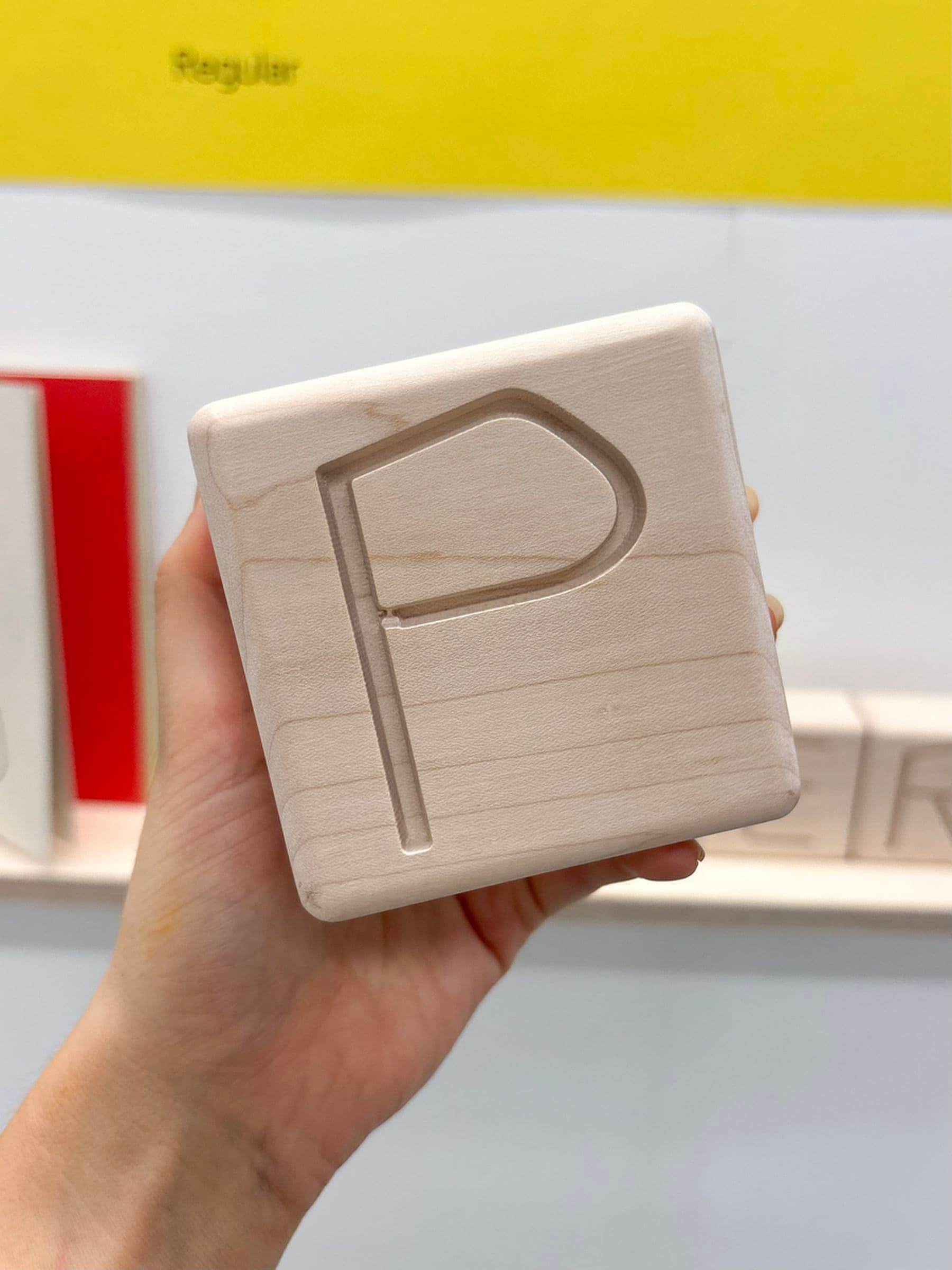 A photograph of a hand holding a wooden block with an engraved letter 'P' against a background with the bottom of a yellow poster and other wooden blocks partially visible in the background.