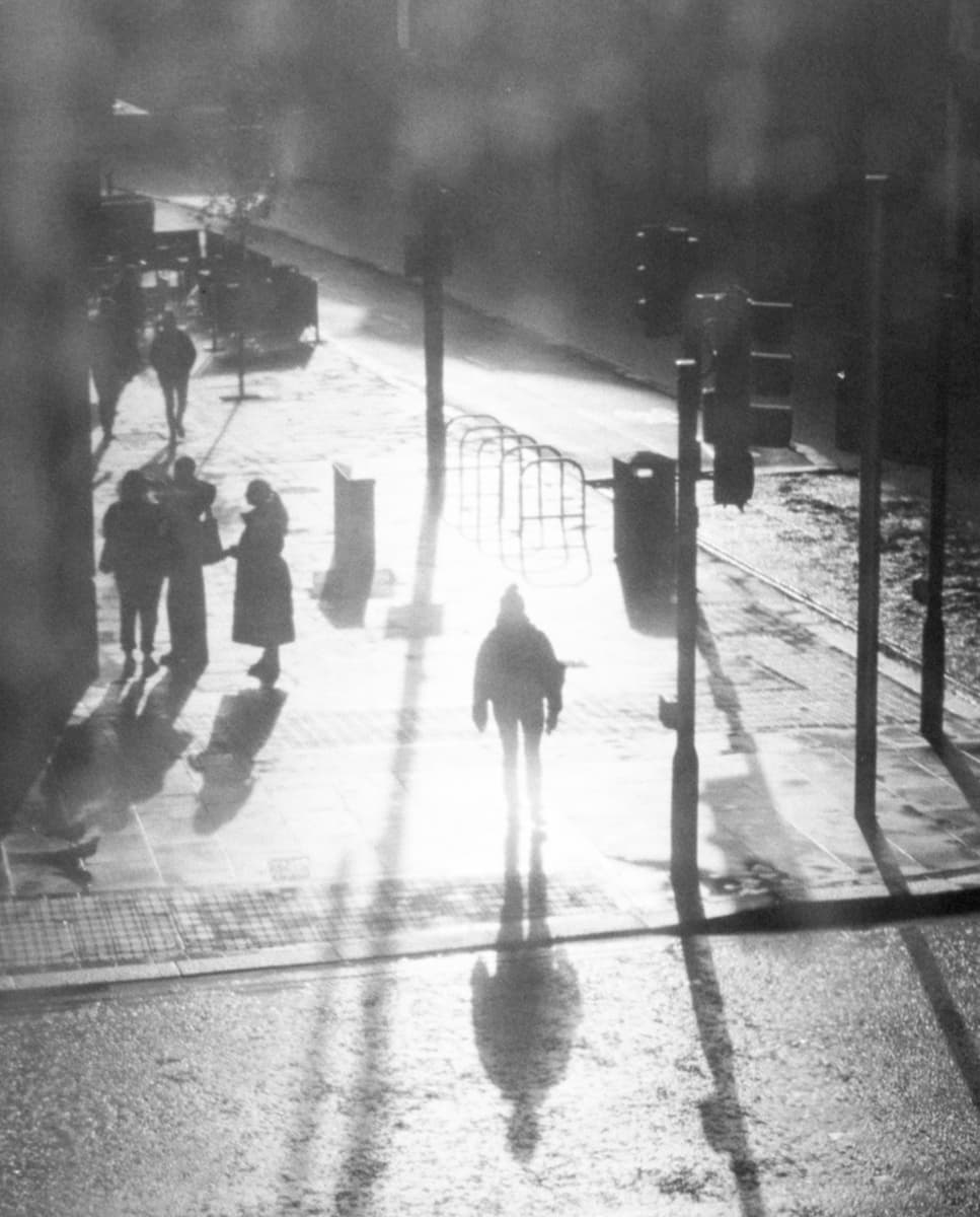 Silhouetted pedestrians walk along a sunlit street, casting long shadows on the wet pavement, with faint figures and traffic lights in the background.