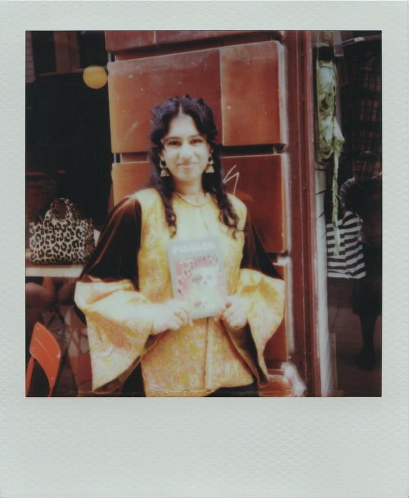 A person in a yellow and brown outfit holds a magazine, standing in front of a brick wall.