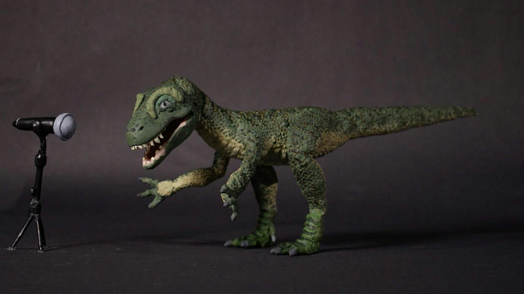 Model of a green dinosaur against a dark background standing in front of a microphone. Its mouth is open as if it is singing.