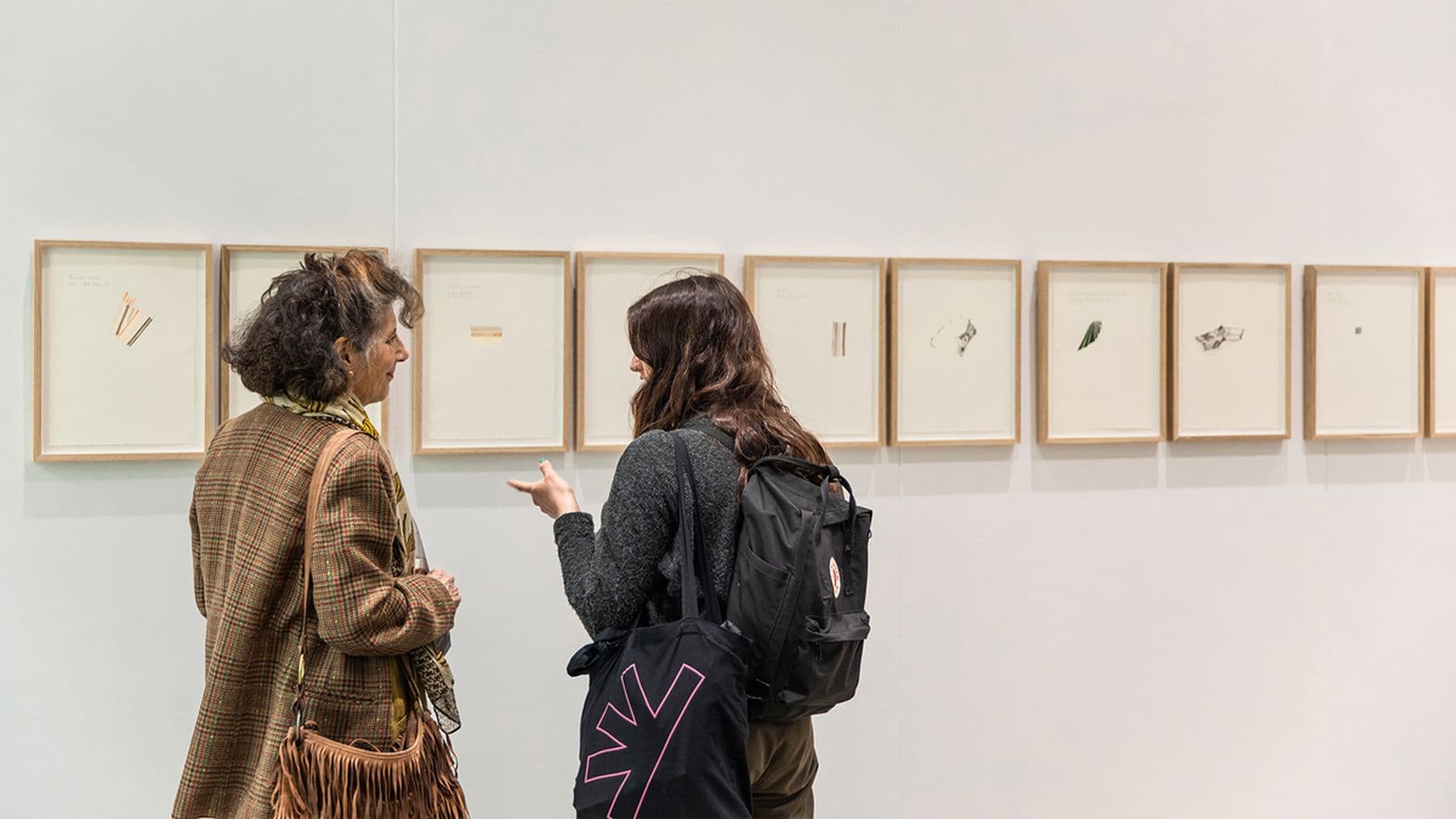 A photograph of a two people talking while viewing a row of small framed minimalist drawings on a white gallery wall.