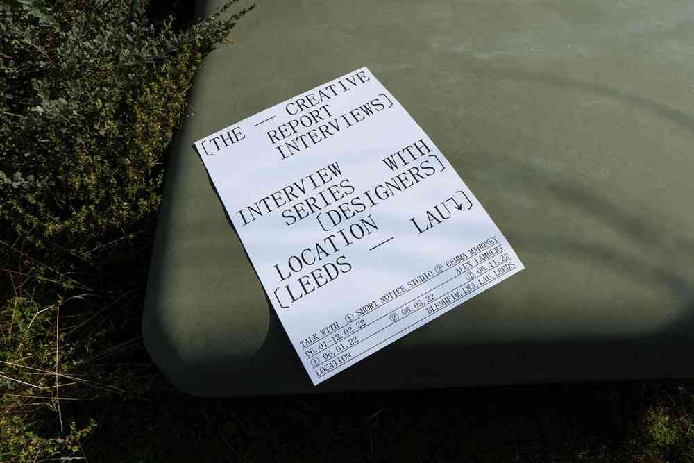 White sheet of paper on a grey stone. The black, uppercase text reads: [The - Creative Report Interviews] Interview series with [designers], location [Leeds - LAU]. There is further text at the bottom of the page that is too small to be read easily.