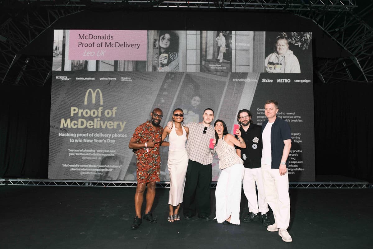 A group of six people smiling and posing on stage in front of a large screen with "Proof of McDelivery" displayed.