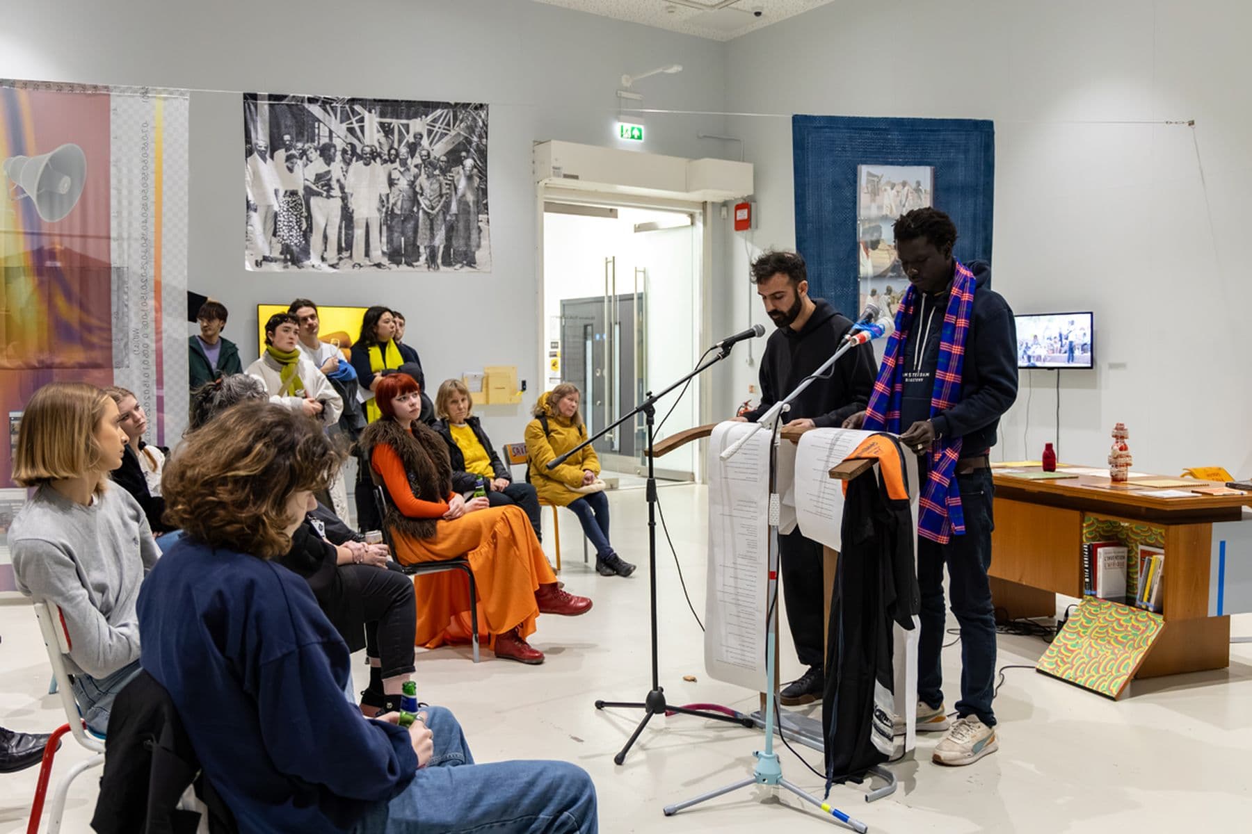 A photograph of two men reading into microphones from long scripts to a seated audience in a gallery with artwork on the walls.