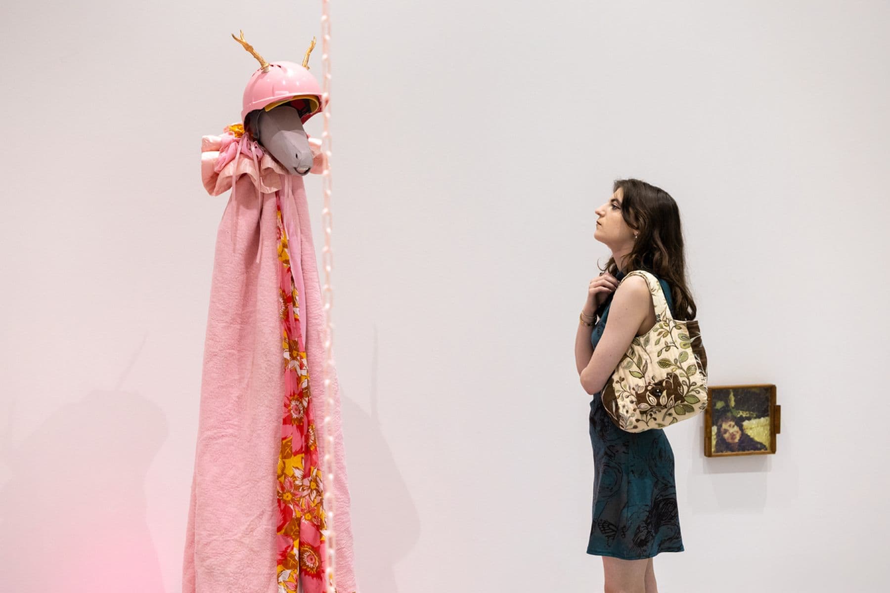 A photograph of a person in dress with a floral tote bag studying a tall pink-draped sculpture wearing a pink helmet with antlers in a gallery.