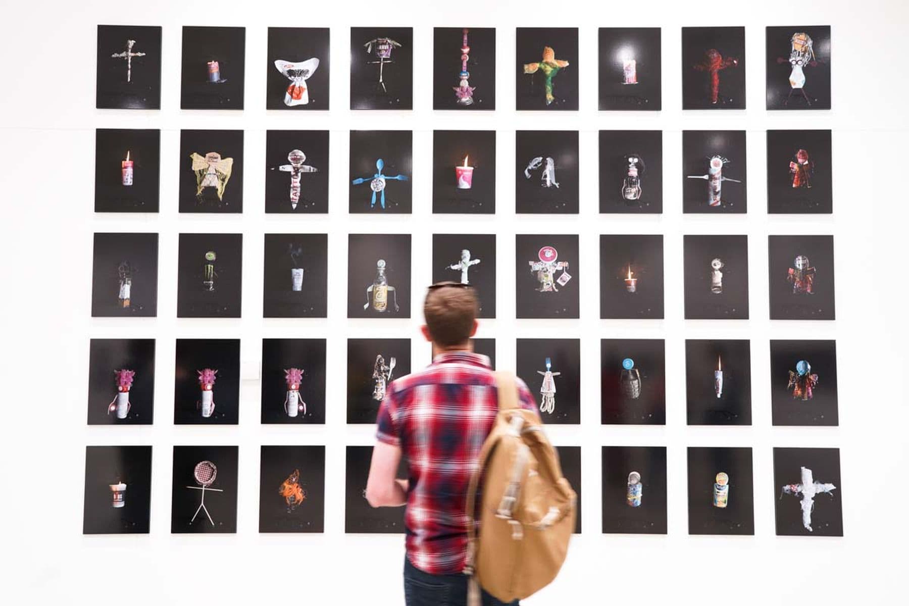 A photograph of a person wearing a backpack viewing a grid of small black-framed photographs displayed on a white gallery wall.