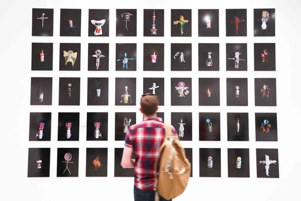 A photograph of a person with their back facing to the camera. They are viewing  multiple photos mounted on the wall of  differing small objects.