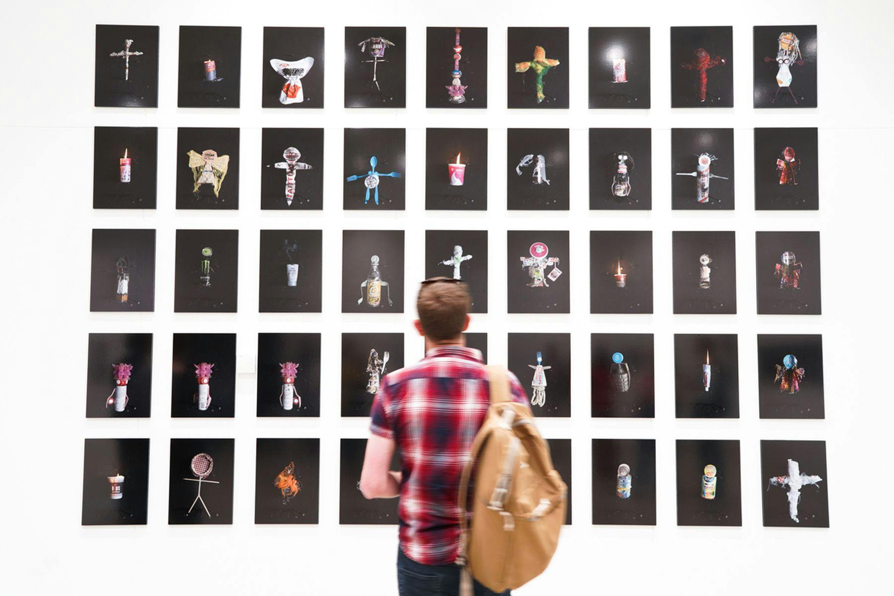 A photograph of a person with their back facing to the camera. They are viewing  multiple photos mounted on the wall of  differing small objects.