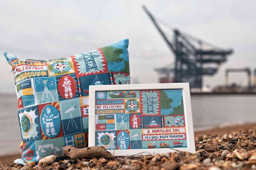 A photograph of a cushion and framed pattern on a pebbled beach with the sea and a crane in the background. The cushion and frame have the same pattern which is blue, red, yellow and green and has astronauts, cranes, globes and computers among other icons.
