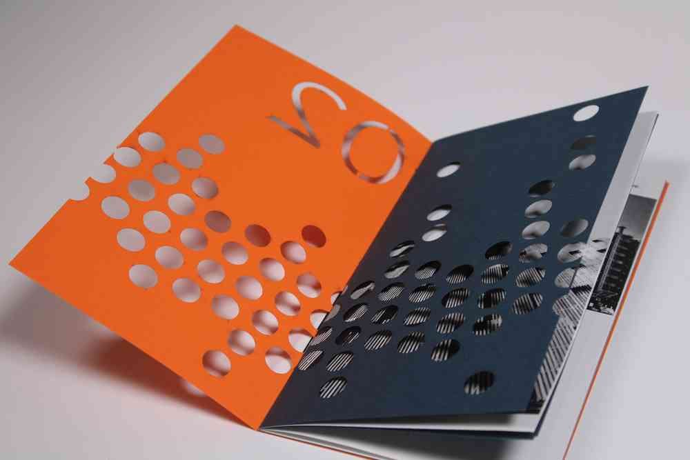 A photograph of an open booklet design, the two opened pages are orange and black. The pages have holes punctured in a linear design and the number 02 on the left-hand page.