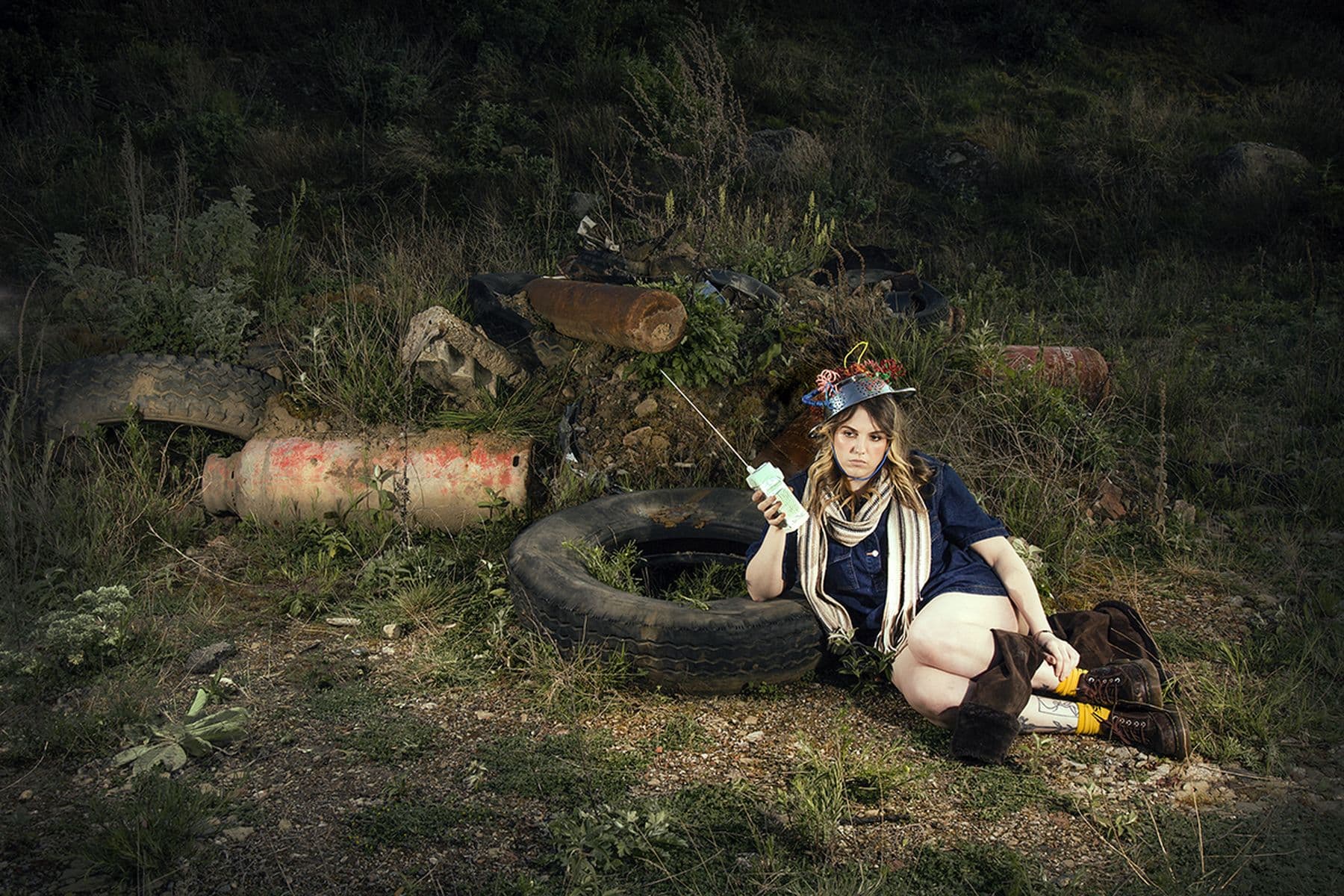 A person sits on the ground in front of a pile of old tires and barrels, wearing a hat and holding a device.