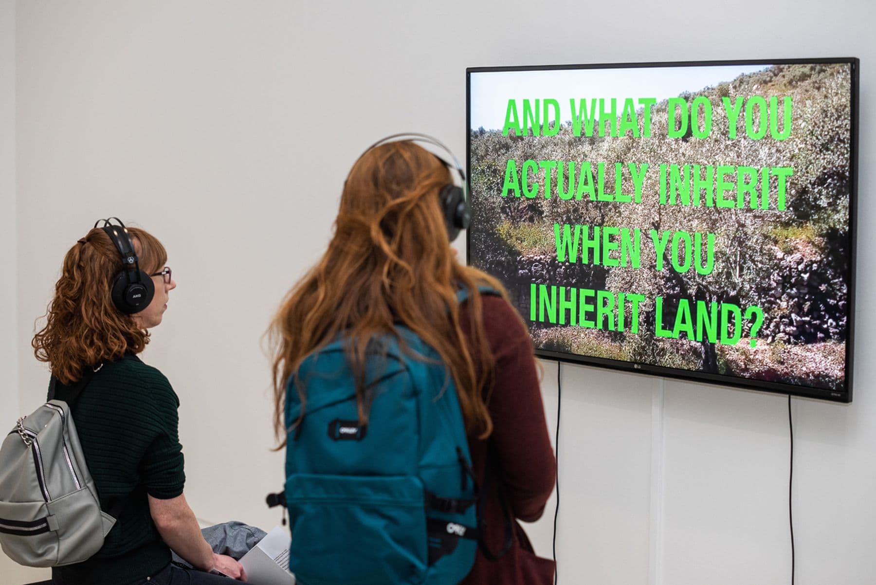 A photograph of two people with headphones watching a wall-mounted screen showing green text that reads: 'And what you actually inherit when you inherit land?'
