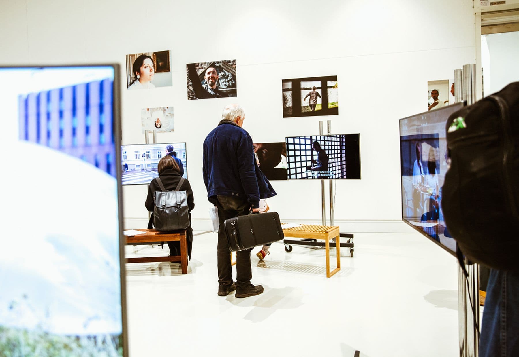 A photograph of people in a bright gallery viewing photos and video screens; a man holding a briefcase stands near wooden benches.