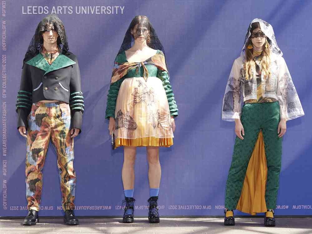 A photograph of three models wearing veils with a lace design. One wears a black suit jacket with a green collar and orange, black and blue patterned trousers. Another wears a green top with a draped scarf and layered orange and white skirt. The third wears a white patterned jacked with green trousers.