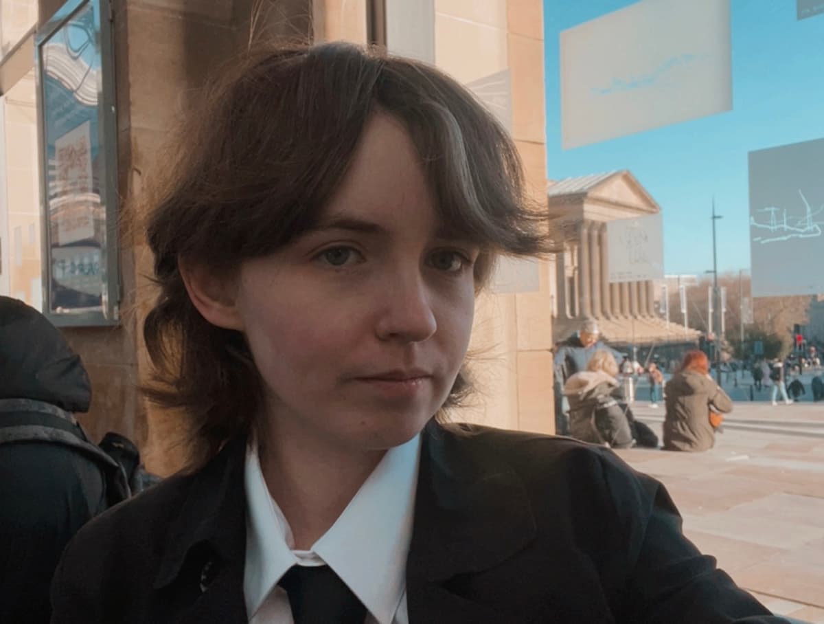A person with short brown hair, wearing a white shirt and black jacket, with people walking in the background.