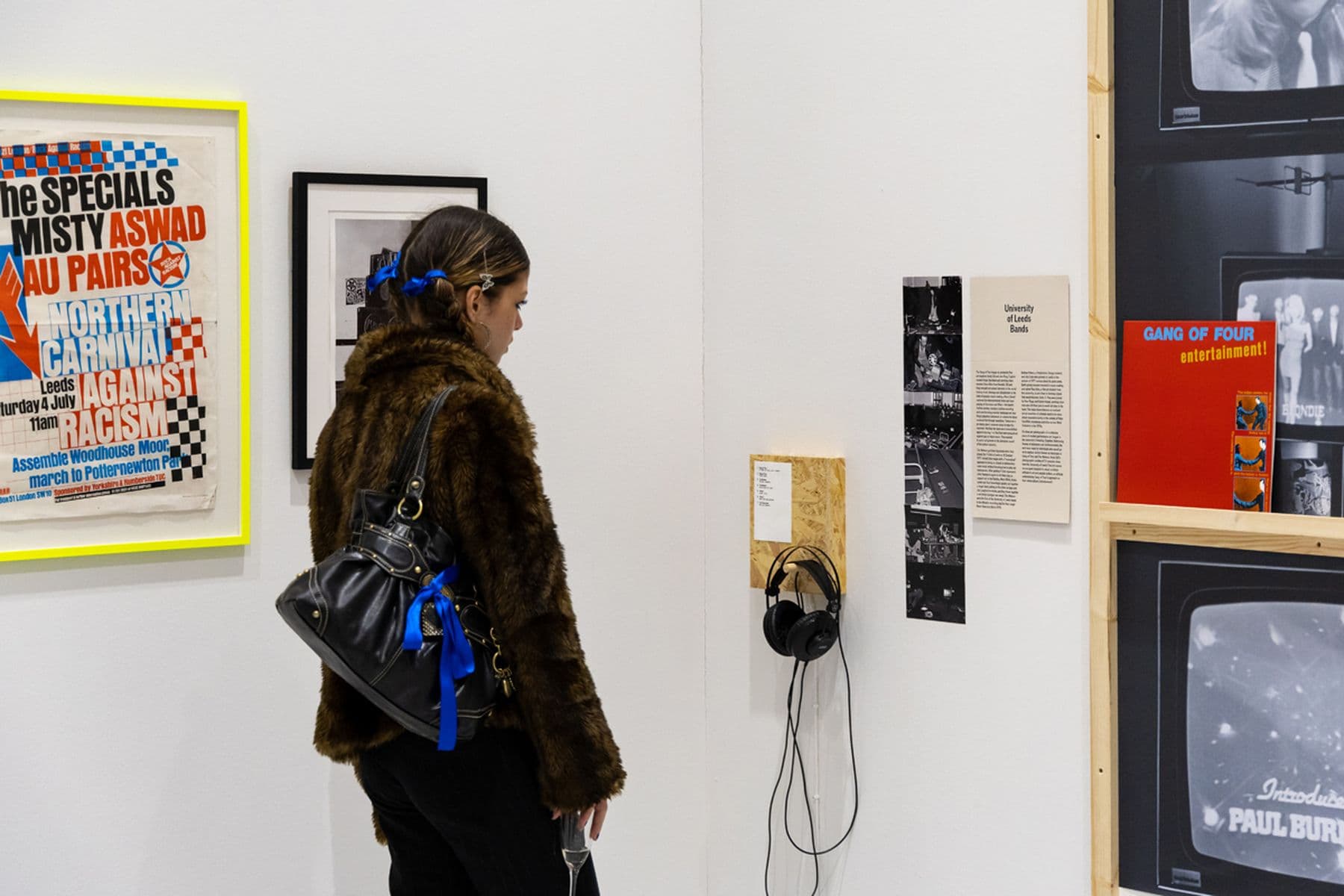 A photograph of a person in a fur coat examining framed posters and wall-mounted headphones in an art gallery.