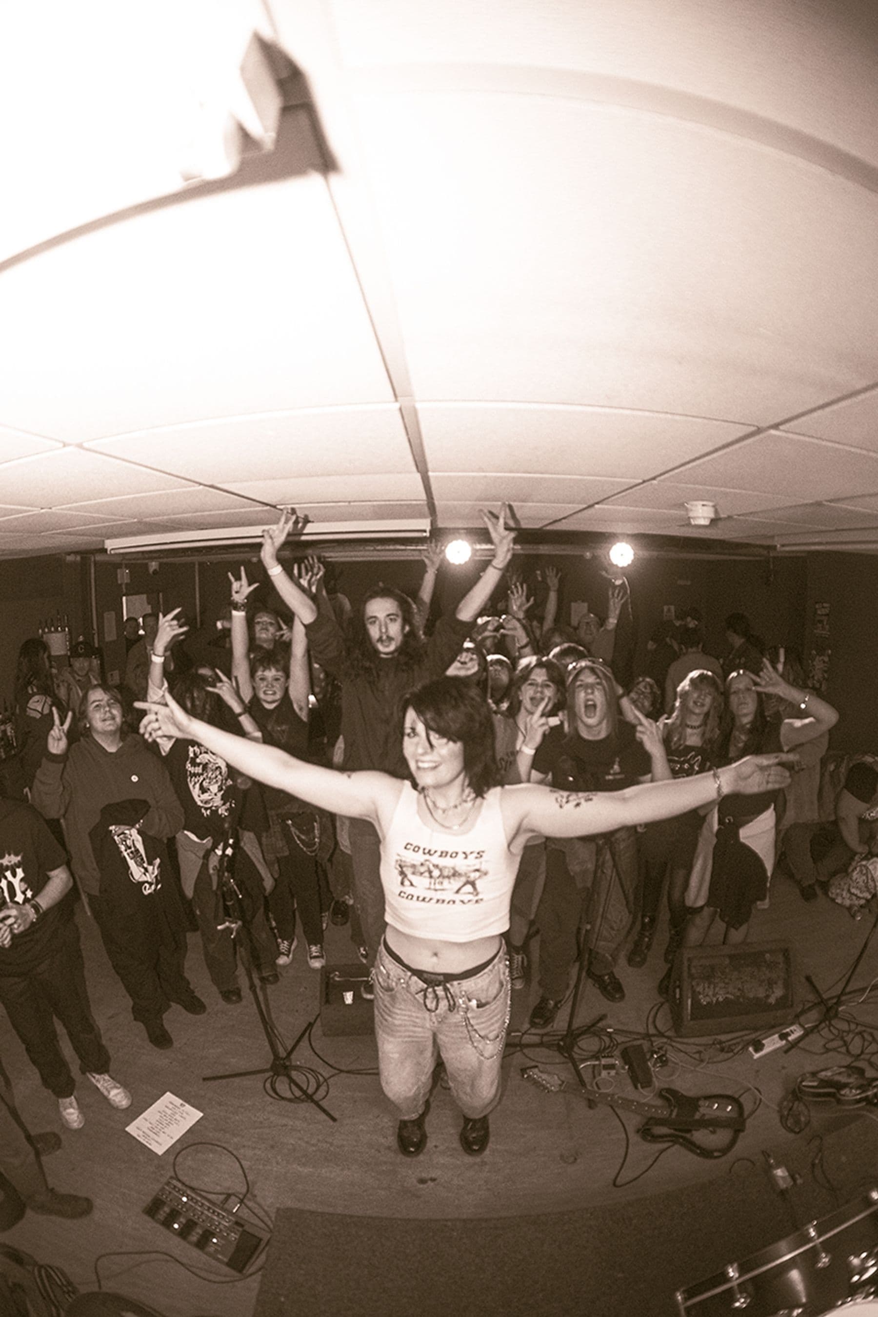 A person stands at the front of a lively, cheering crowd with arms outstretched in a small indoor venue, surrounded by musical equipment.