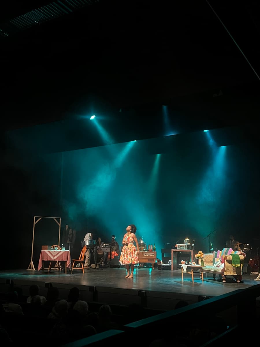 A person in a floral dress performs on a stage against a blue spotlight. The set is dressed to look like a living room, with a sofa and dining table.