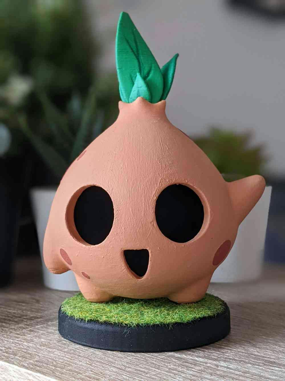 A photograph of a terracotta clay character that has a large rounded face with a smiling expression, there is a green plant emerging from the top of their head. The character is waving and standing on a round grass step.
