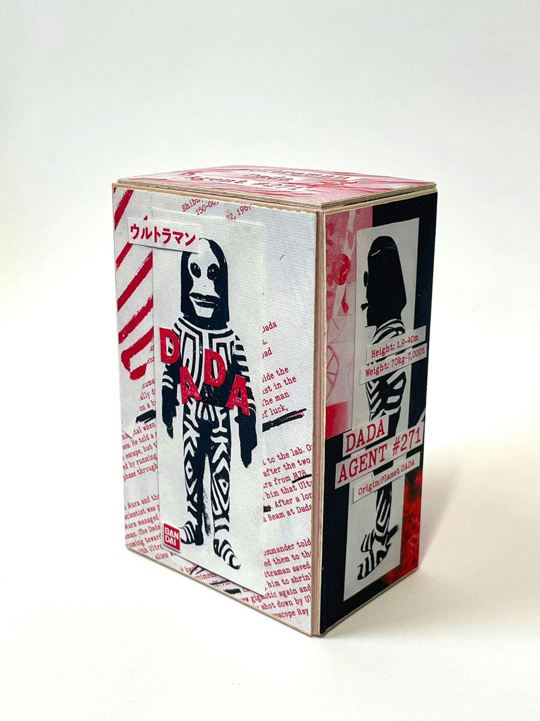A photograph of a cardboard box with a black alien like figure positioned behind the title in pink lettering “dada”. There is another title in Japanese and paragraphs of pink text.