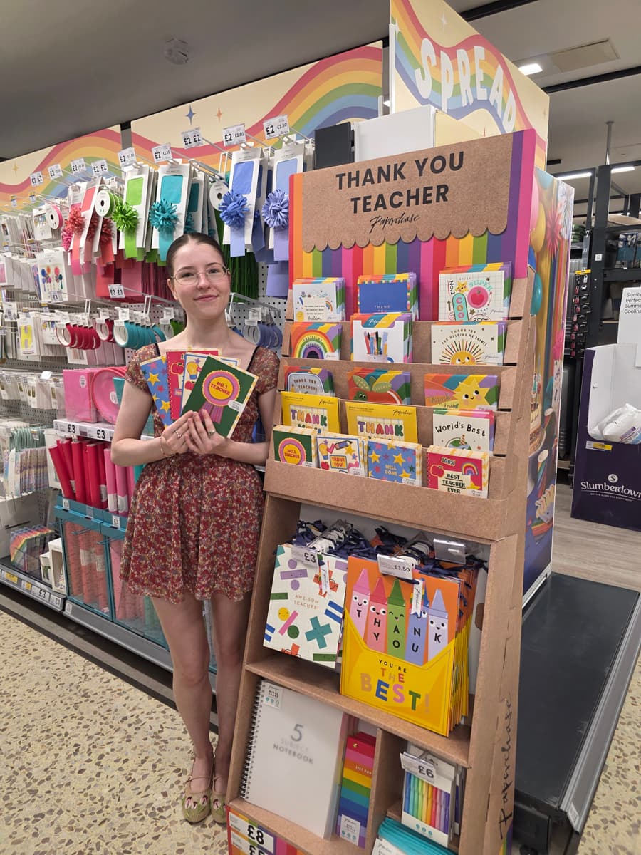 A person stands in a store next to a display of colorful "Thank You Teacher" cards and gifts, holding several cards, with rainbow decorations above.