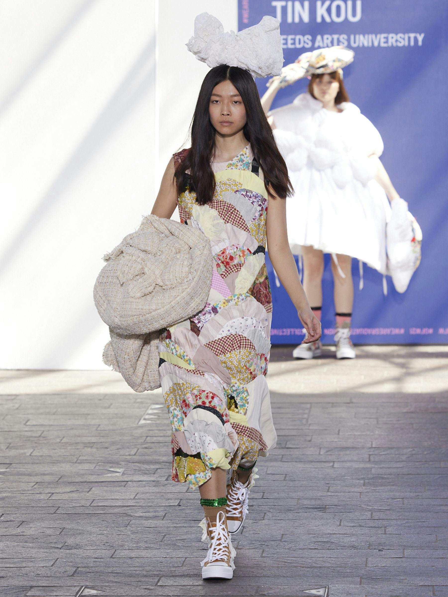 A photograph of a model walking down a runway wearing a long sleeveless patchwork dress. The patchwork consists of floral designs in pastel colours such as pinks, creams, blues, yellows and crimson. There is a large tweed flower adornment on the left side of the dress. The model wears a large bow on the top of their head and dark yellow sneakers.