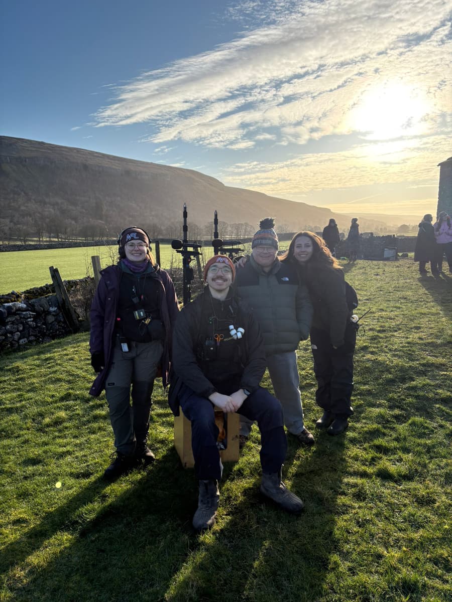 A group of four people in outdoor attire smile at the camera in a grassy field with a scenic mountain backdrop and a bright sunlit sky.